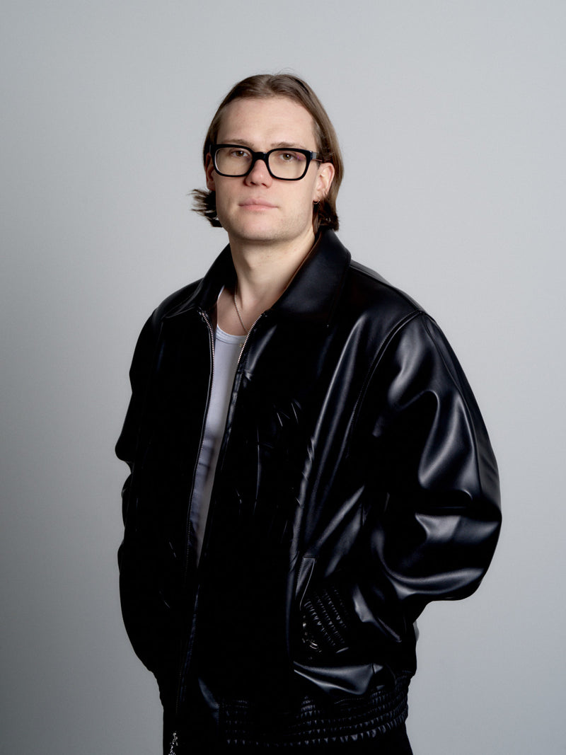 Person wearing a deadHYPE Vegan Leather Jacket (Black)  and glasses against a gray background